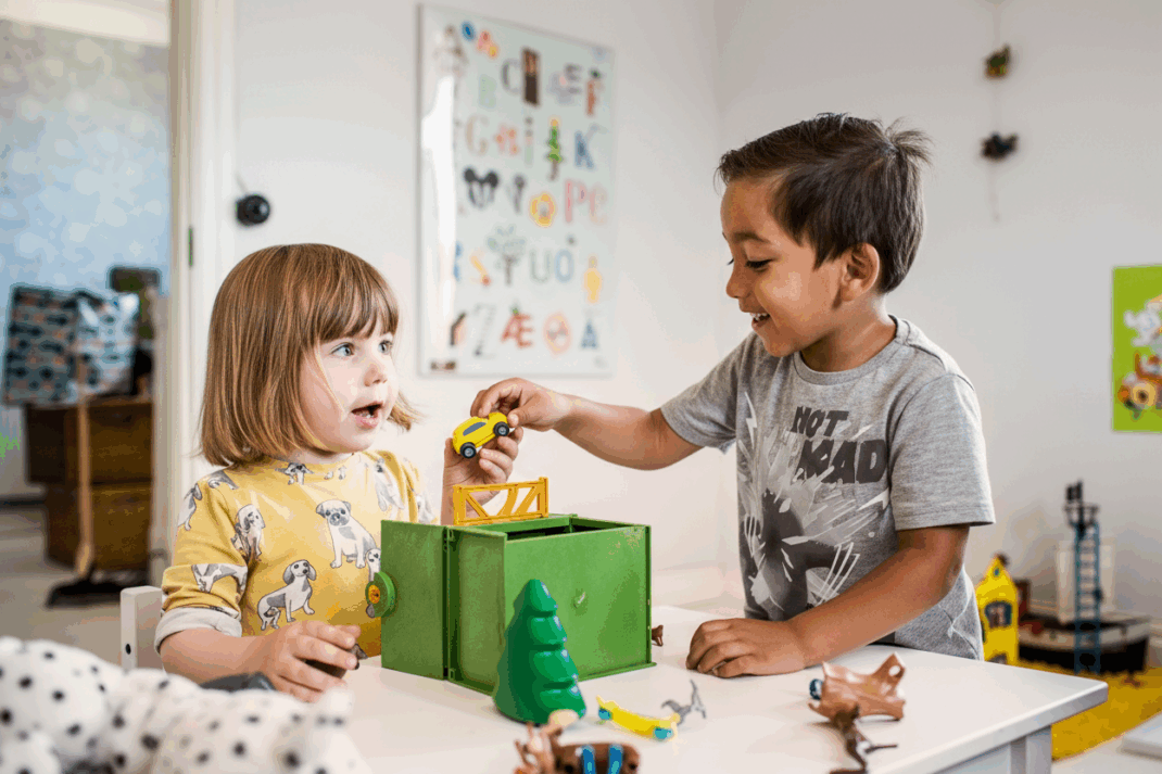 Two young children play with toy cars and animal figures at a table in a brightly decorated room with alphabet art on the wall.